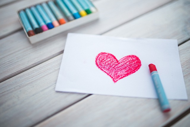 A box multiple color pastel crayons and a red heart drawn by a red pastel crayon on a white piece of paper